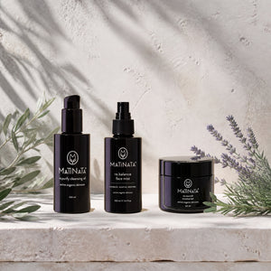 Three black MATINATA skincare bottles and a jar arranged on a light stone surface with olive branches and lavender sprigs against a textured off-white wall with soft leaf shadows.