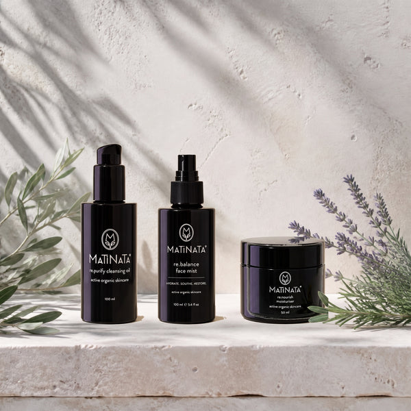 Three black MATINATA skincare bottles and a jar arranged on a light stone surface with olive branches and lavender sprigs against a textured off-white wall with soft leaf shadows.