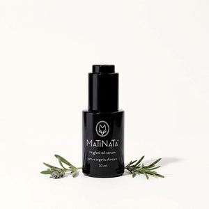 A 30 ml black bottle of MATINATA re.glow Oil Serum, styled with rosemary and lavender sprigs on a clean white background.