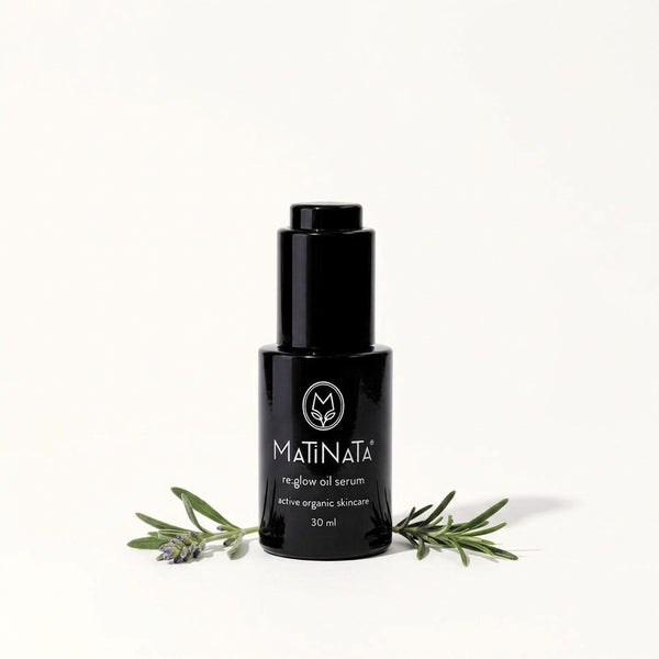 A 30 ml black bottle of MATINATA re.glow Oil Serum, styled with rosemary and lavender sprigs on a clean white background.