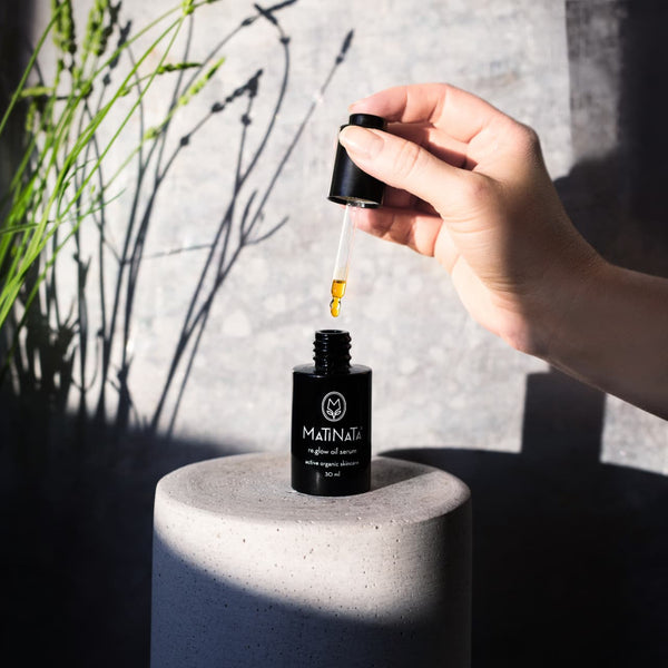 A hand holds an open dropper bottle above a black cylindrical pedestal, with a single drop of golden oil dripping between the glass pipette and the bottle. The bottle label reads MATINATA.