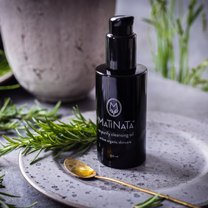 A bottle of MATINATA cleansing oil stands on a speckled gray plate with a gold spoon, surrounded by rosemary sprigs and a blurred stone vessel in the background.